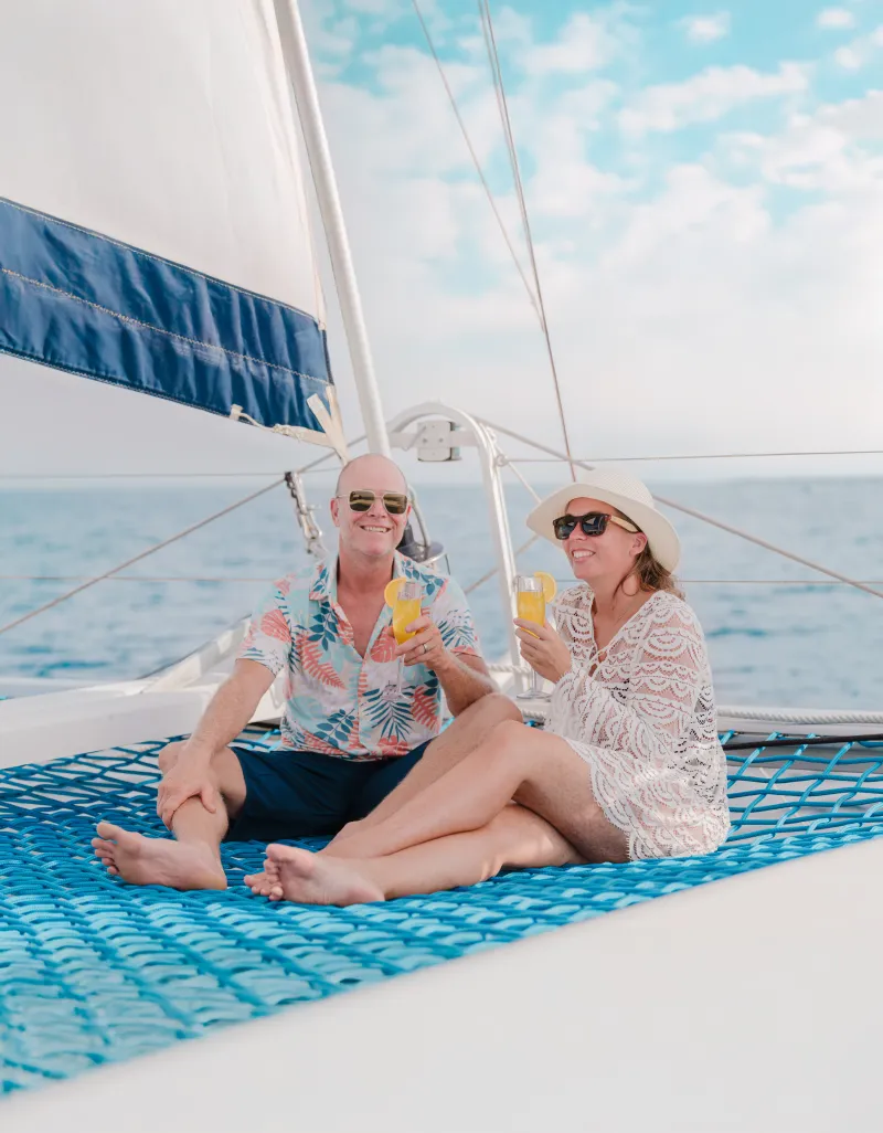 Couple on sailboat net, wearing sunglasses and holding drinks, under blue sky.